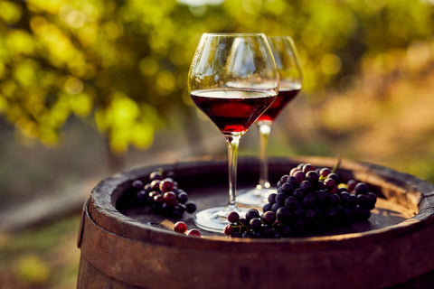 Best red wines displayed in glasses with fresh grapes on a wooden barrel in a vineyard setting.