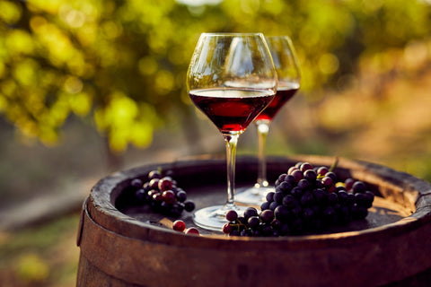 Best red wines displayed in glasses with fresh grapes on a wooden barrel in a vineyard setting.