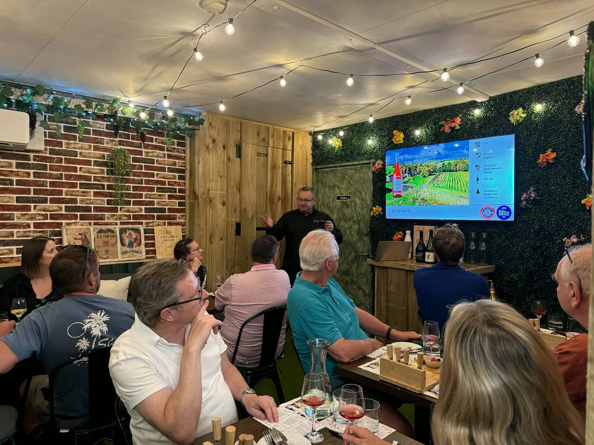 Jamie hosting a tasting in the snug, guests at long tables with glasses of rosé, the wine on the screen behind him